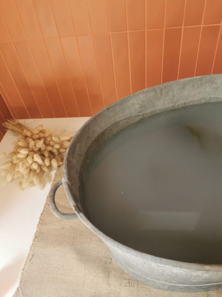 Salle de bain moderne avec carrelage mural ocre. Au sol une grande baignoire en zing contenant de l'eau savonneuse